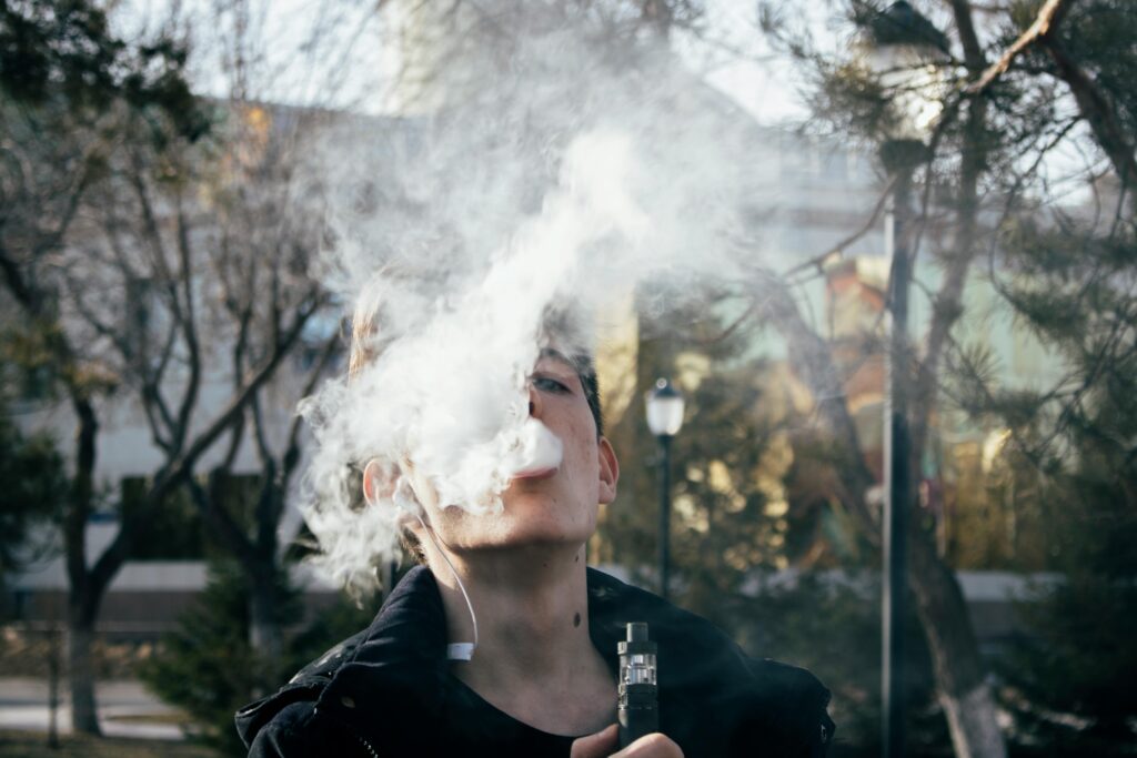 Foger vapes Portrait of a young man vaping in a park, releasing clouds of vapor on a clear day.